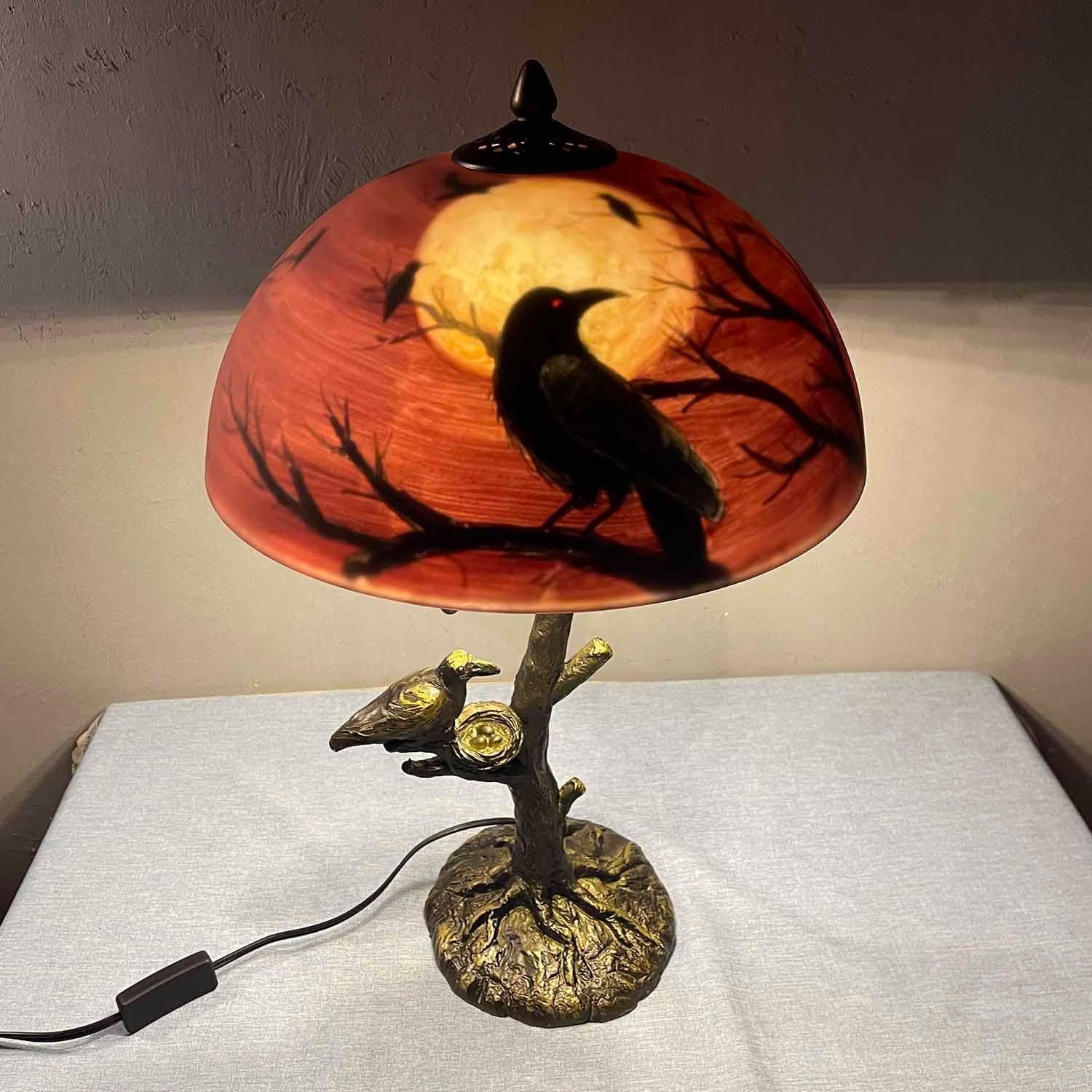 Stained glass table lamp with a raven silhouetted against a full moon, on a bronze tree-trunk base with a second raven and nest