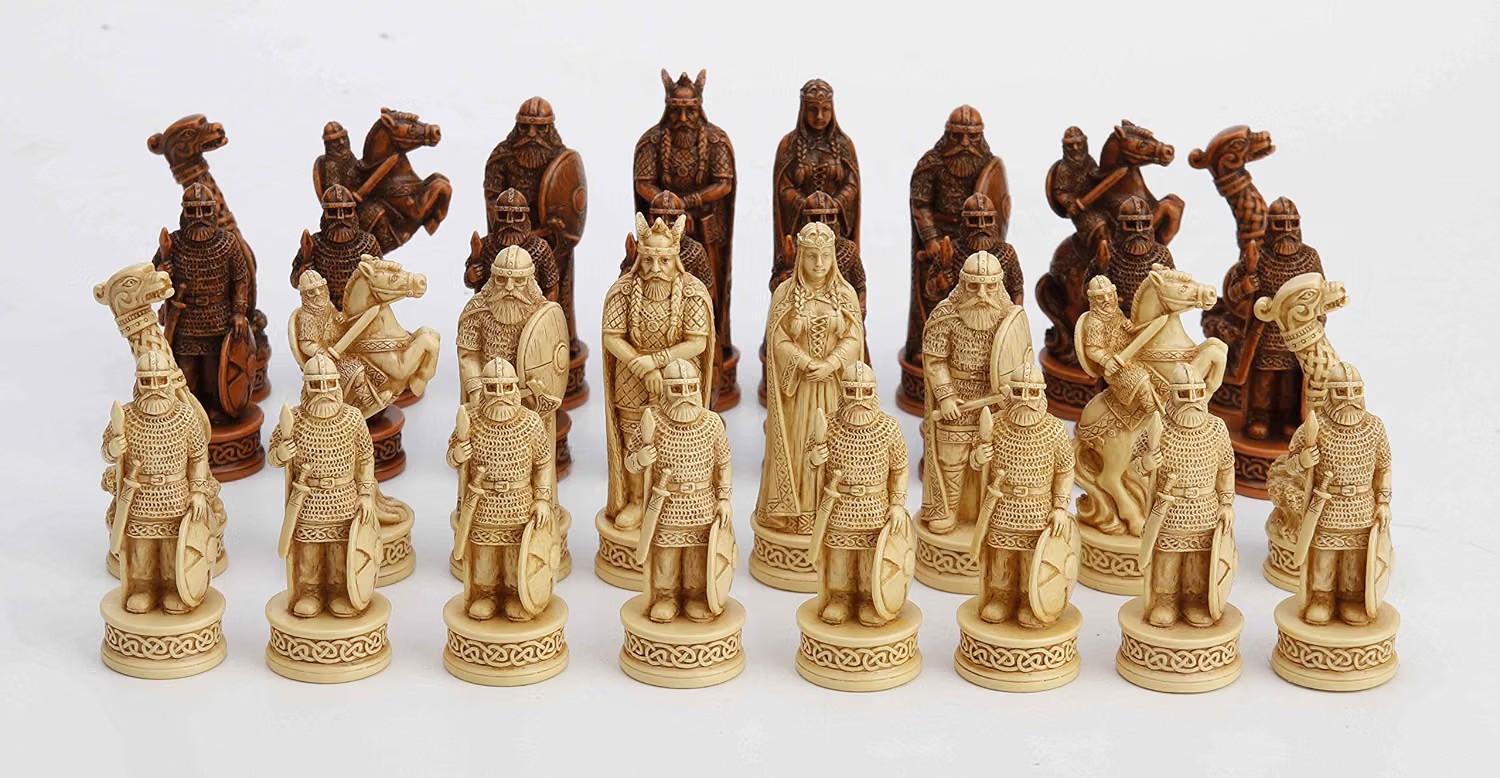 Isle of Lewis-style Viking chess set with full-figure shield-bearing warriors, mounted riders, a king and queen, and dragon-prow ship rooks, in cream and dark bronze finishes with Celtic-knot carved bases