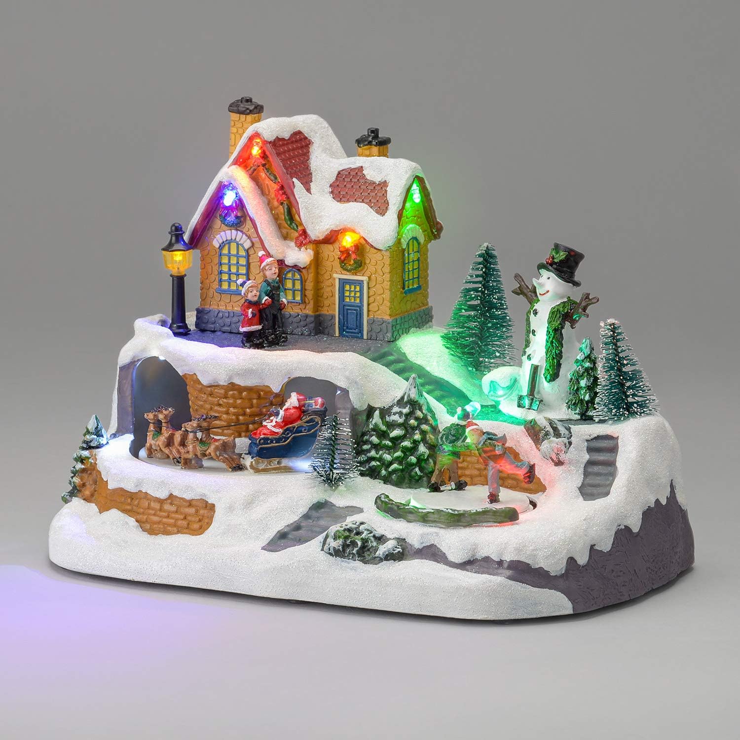Hand-painted Christmas village scene with a snow-covered cottage lit in multicoloured LEDs, a snowman, skaters on a frozen pond, and Santa's sleigh and reindeer passing through a tunnel