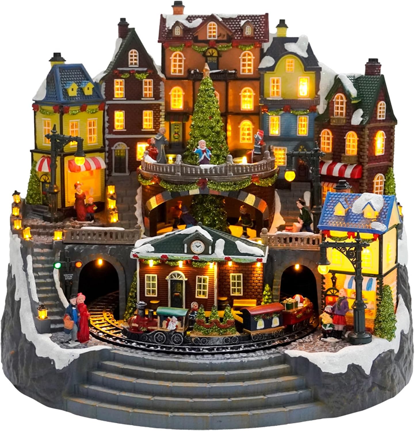 Large tiered Christmas town display with lit townhouses and shopfronts around a tall Christmas tree, a clock-tower station below, and a little train running through stone arches past villagers and stone steps