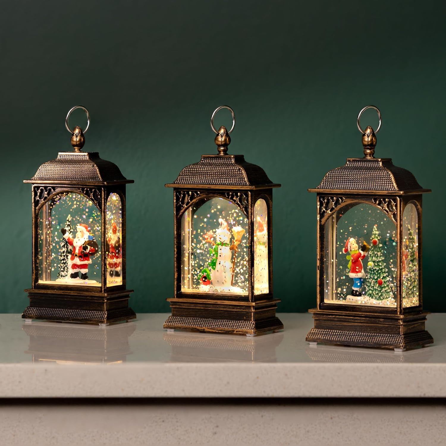 Three bronze-effect light-up snow globe lanterns on a shelf, holding glittering Santa, snowman, and Christmas tree scenes