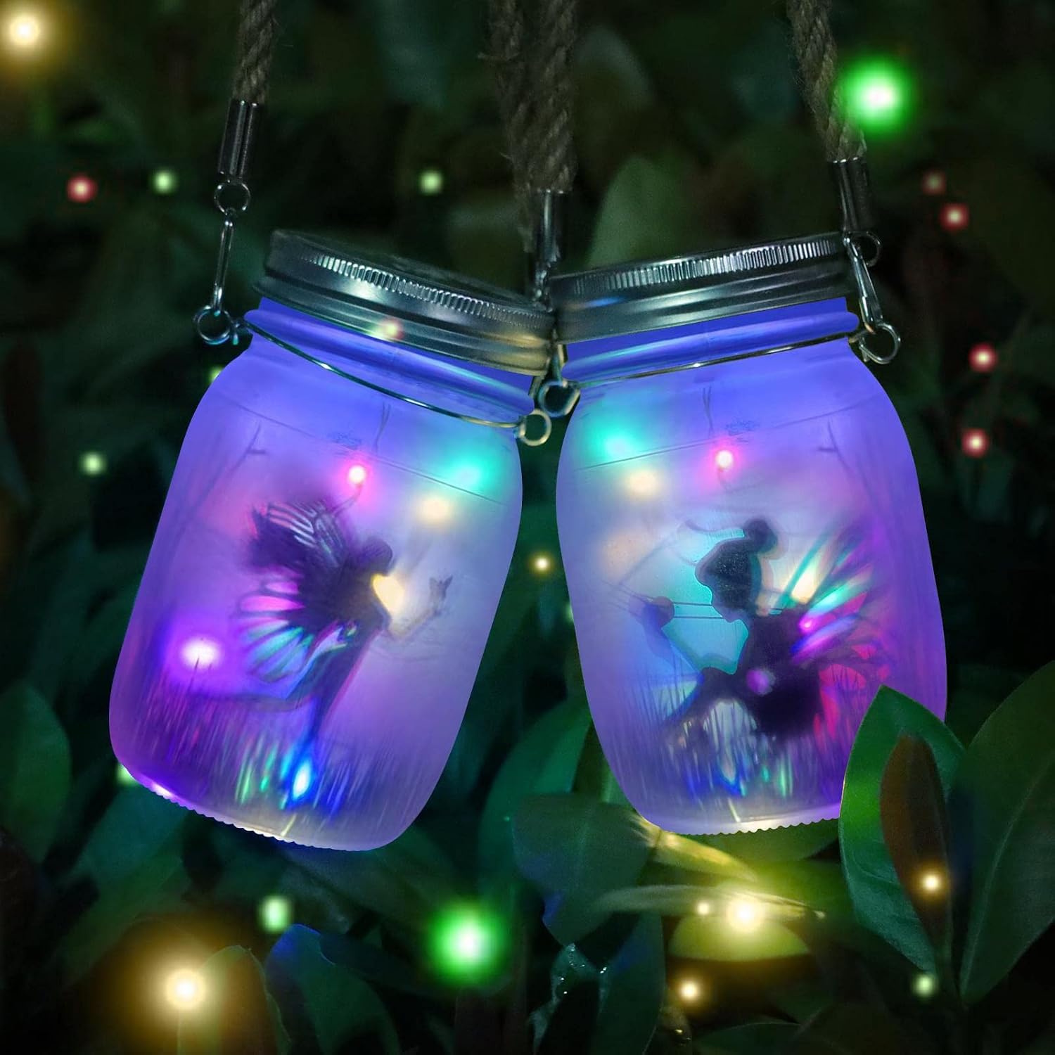 Two frosted mason jar lanterns hung on rope among leaves at night, each glowing purple and blue with multicoloured fairy lights and a winged fairy silhouette inside